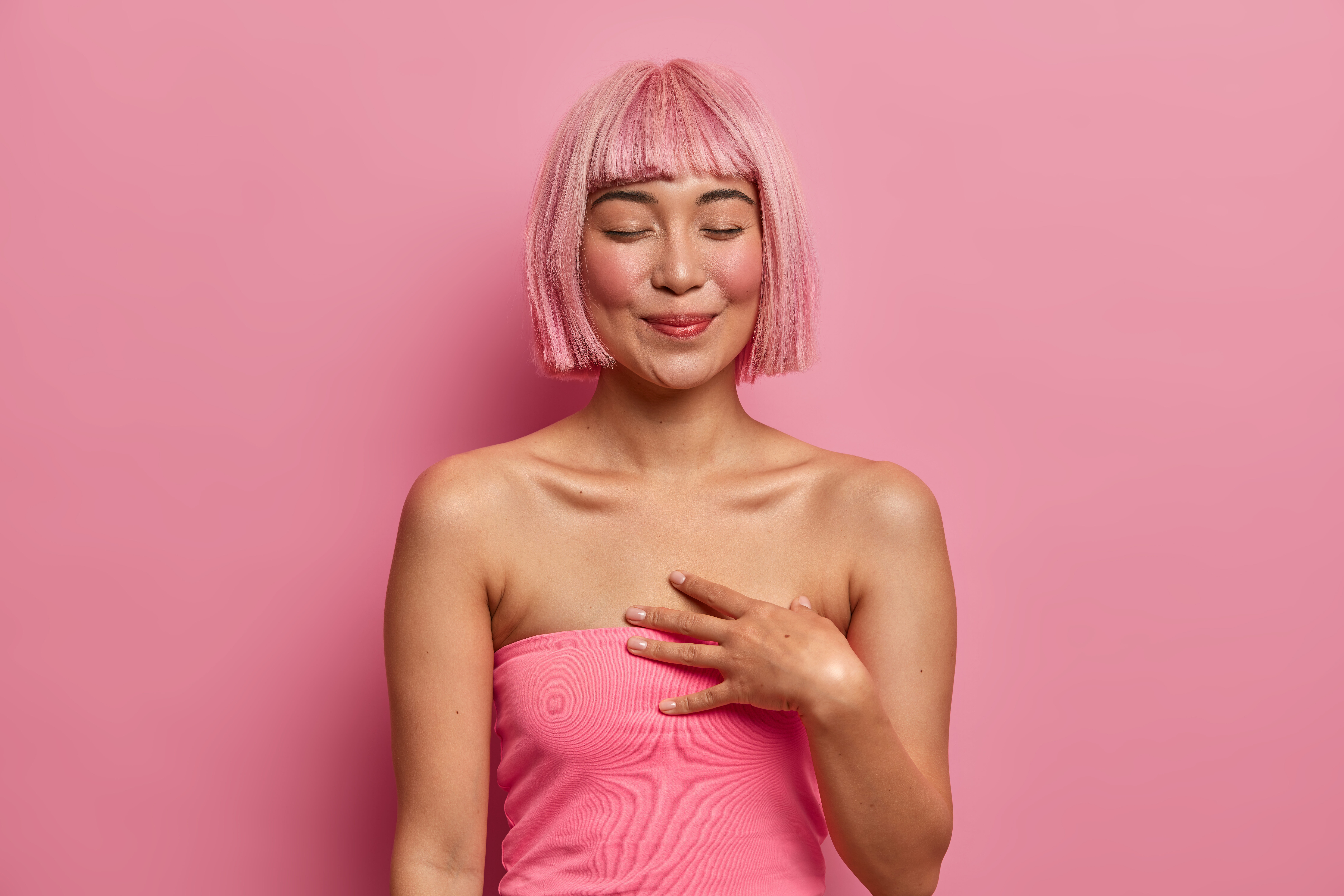 Tender touched woman closes eyes keeps hand near heart happy receive gift recalls lovely date has dyed pink hair dressed casually 1
